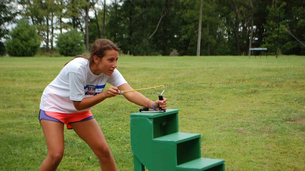 Young woman aiming a wrist rocket/slingshot outdoors.